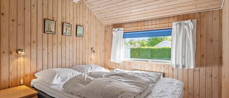 A bedroom with wooden walls, a bed with white bedding, a window with curtains, and a skylight.