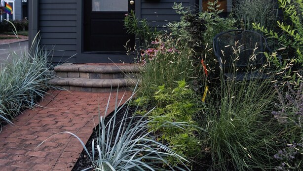 A modern two-story house with a black front door, a small porch, and a well-maintained garden with various plants and a brick pathway. - Short North, Free Parking, Italian Village (Columbus)