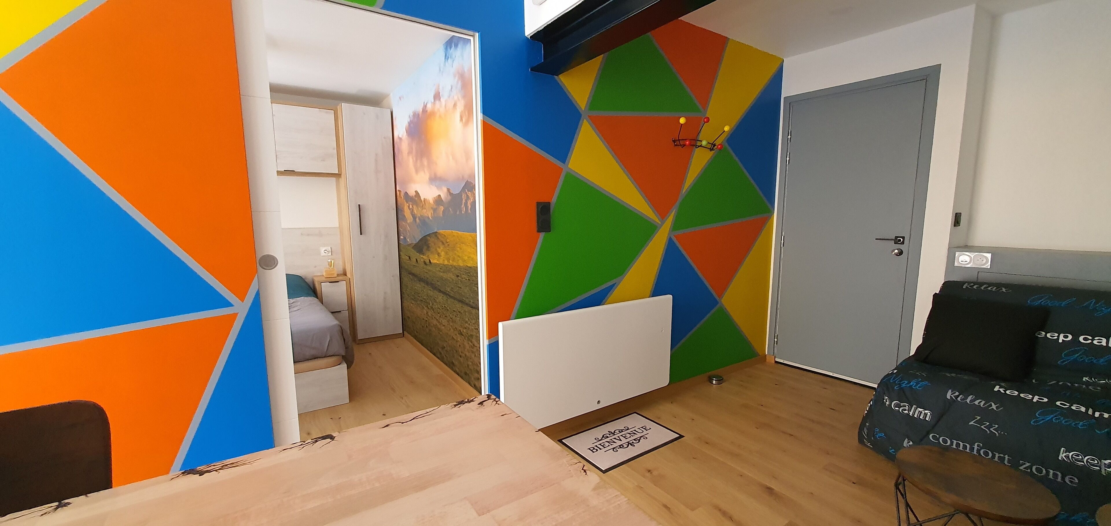 Ett rum med en färgstark, geometrisk väggdesign, en säng, ett skrivbord och en stol med ett tema som uppmanar till lugn.