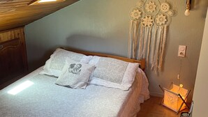Un dormitorio con techo inclinado, una cama con ropa blanca, un atrapasueños en la pared y una mesita de noche con una lámpara.