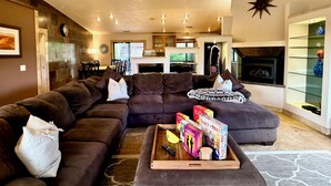 A living room with a sectional sofa, a coffee table with board games, a fireplace, and a kitchen area in the background. - Serene Escape in West Sedona w/ Gorgeous Views (Sedona)