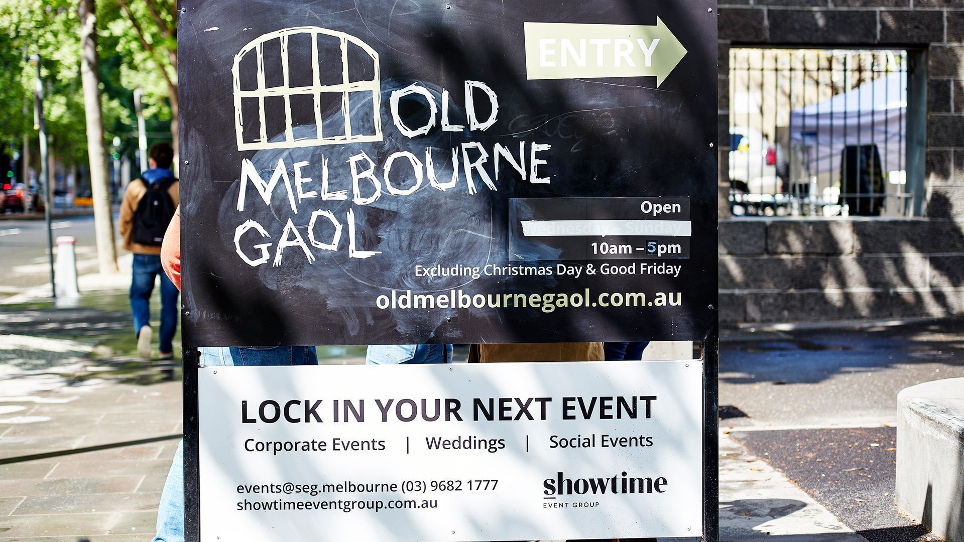 Een bord voor Old Melbourne Gaol met een pijl die de ingang aanwijst, openingstijden en een websiteadres.