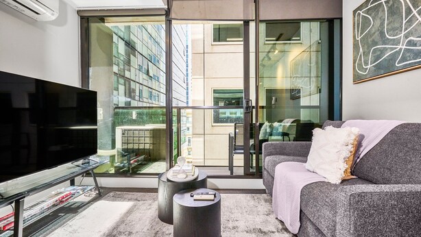 A modern living room with a large flat-screen TV, a grey sofa with a throw blanket, a coffee table, and a glass railing overlooking a cityscape. - Melbourne CBD Stay - Balcony, Gym & Prime City Location (Melbourne)