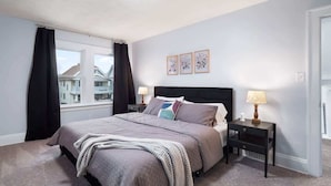 Une chambre à coucher avec un grand lit, deux tables de chevet, une fenêtre avec des rideaux et une vue sur les bâtiments environnants.