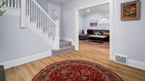 Un vaste espace de vie doté d’un plancher en bois, d’un tapis à motifs rouges, d’un canapé gris et d’un escalier avec des balustrades blanches.