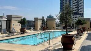 Une piscine entourée de chaises longues et de plantes en pot, avec des bâtiments modernes en arrière-plan et un ciel dégagé.