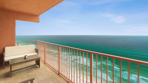A balcony with a white bench and a table, overlooking a beach with clear turquoise water and a clear sky.