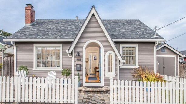 A two-story house with a grey siding exterior, a central entrance with an archway, and a white picket fence. - Peaceful, Updated Cottage. Walk to Town & Beach. (Pacific Grove)