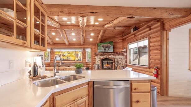 A modern kitchen with wooden cabinetry, a central island with a dishwasher, a stone fireplace, and a large window with a view of autumn trees. - Buffalo Lodge|Hot Tub, EV Charge, Gated, By Slopes (Big Bear)