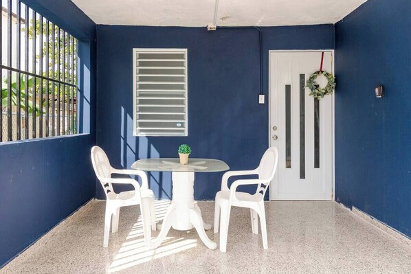 A small, enclosed patio area with a round table and four white chairs, a door with a wreath, and a window with blinds. - Charming 2-bedroom house in Vega Baja with WiFi and AC, Right Near the Beach (Los Naranjos)