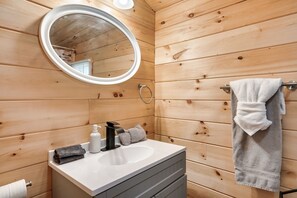 A wooden-paneled bathroom with a round mirror, a sink, a towel rack, and a towel. - Amazing Location - Secluded/Private - Cozy 2-bedroom Log Cabin - Hot Tub (Sevierville)