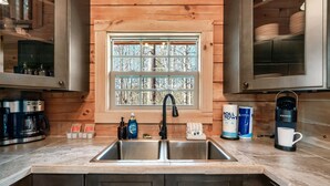 Une cuisine équipée d’un évier à deux bacs, de meubles en bois, d’une fenêtre donnant sur les arbres, ainsi que de divers ustensiles de cuisine, comme une machine à café, du liquide vaisselle et une tasse.