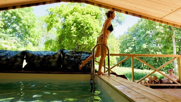 Una terraza de madera que da a una piscina con un área cubierta y árboles al fondo.