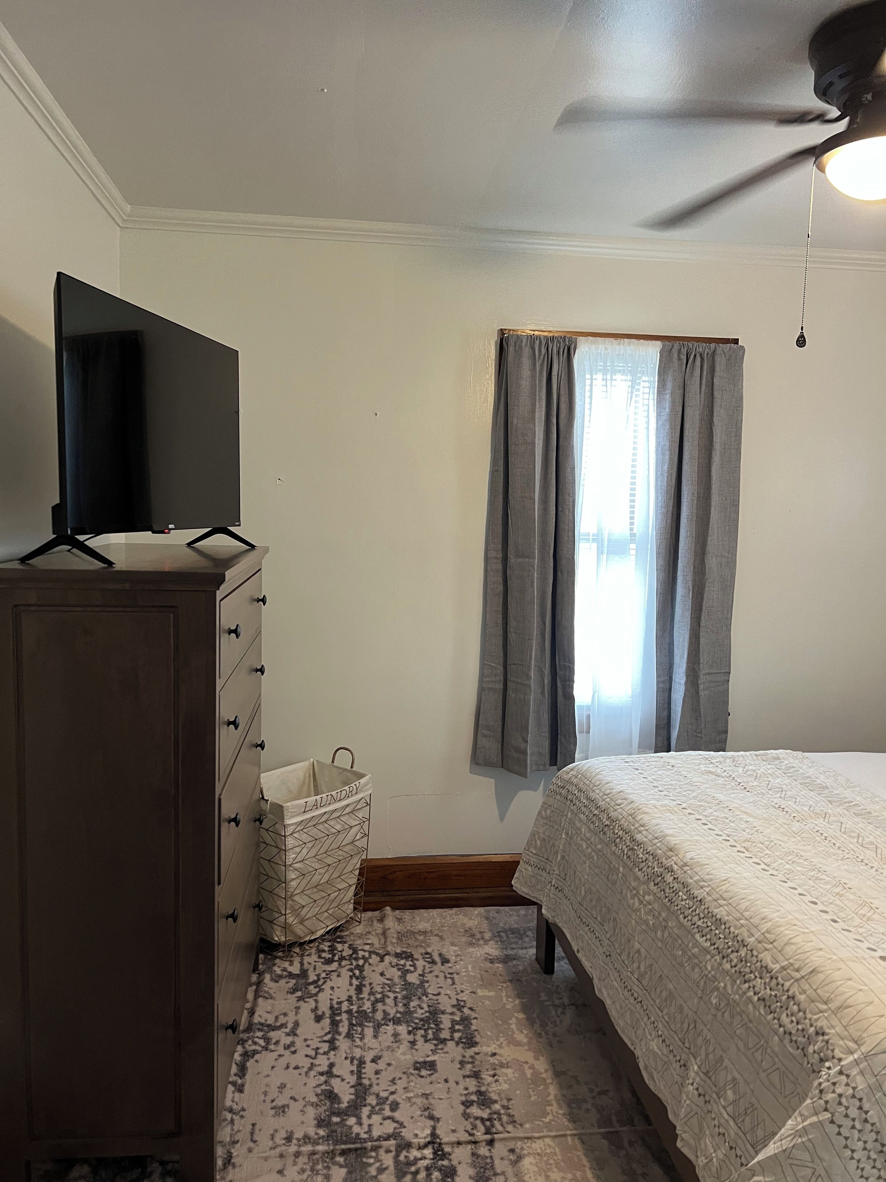 Un dormitorio con cama, cómoda, televisor encima y ventana con cortinas, además de un ventilador de techo.