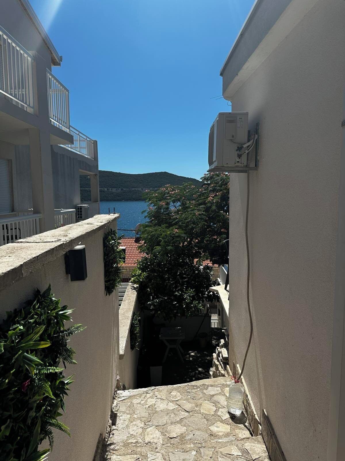 Eine schmale Steintreppe führt zu einem Balkon mit Blick auf ein Gewässer und eine entfernte Hügelkette.
