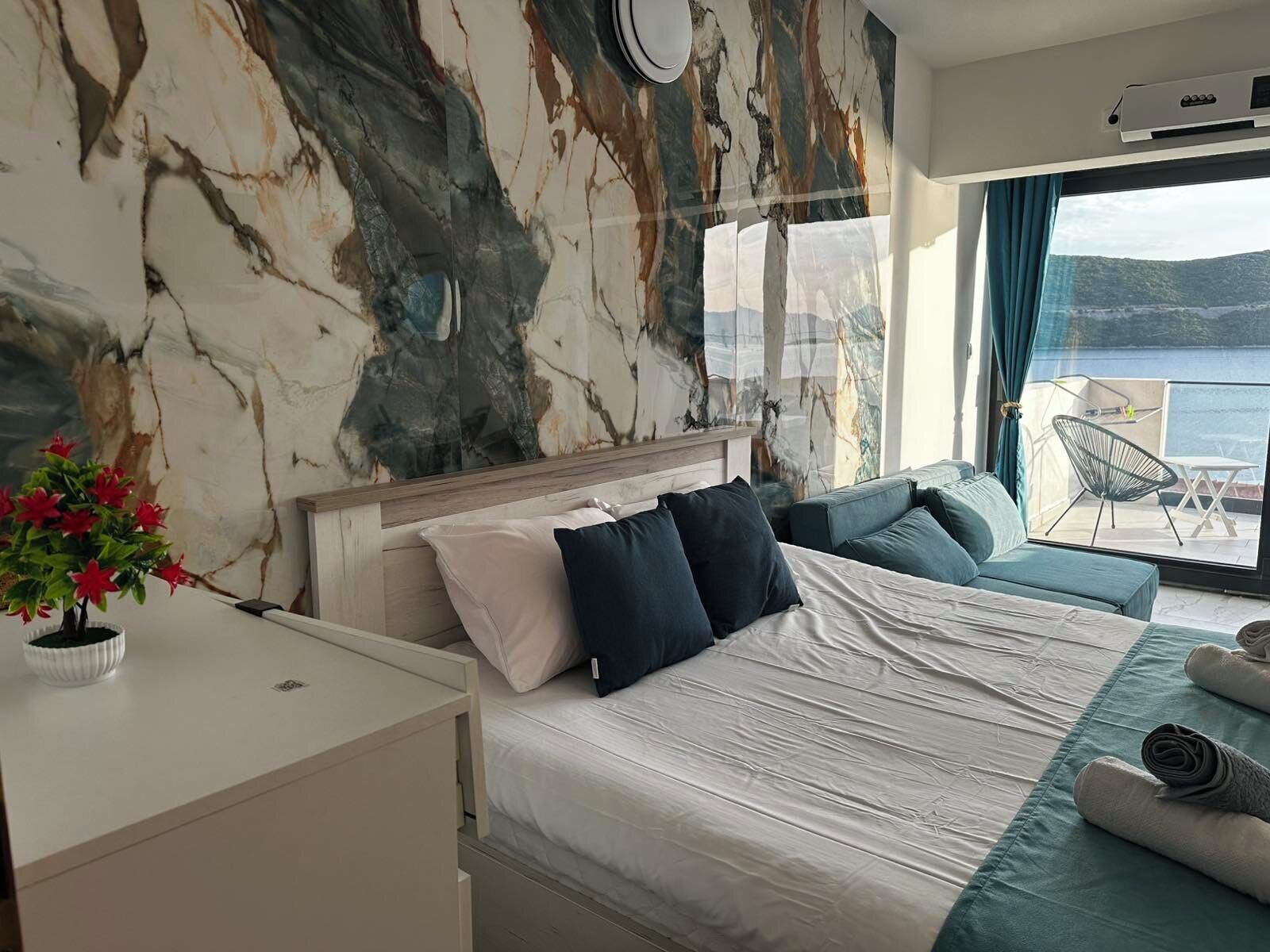 A bedroom with a bed, a desk, a potted plant, and a view of the sea.