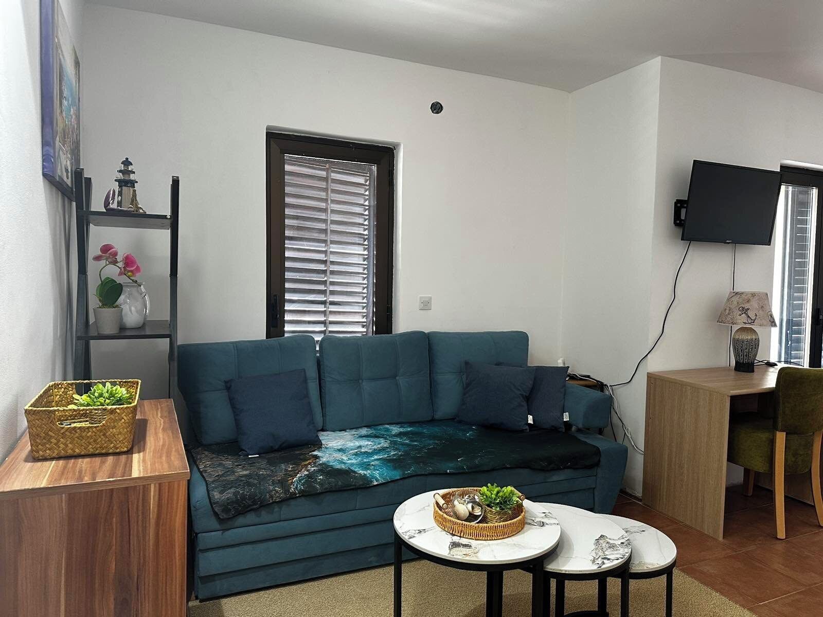 A modern living room with a teal sofa, a small round table, a TV, and a window with blinds.