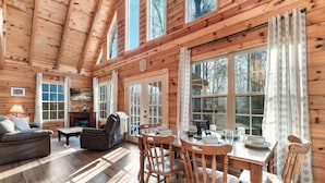 A cozy cabin interior with wooden walls and floors, a dining table set with chairs, a fireplace, and large windows. - Log Cabin Amazing Location - Private - Cozy 3-bedroom Sleeps 8 - Hot Tub (Sevierville)