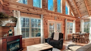 A log cabin interior with a fireplace, a sofa, a dining table, and large windows offering views of the forest. - Log Cabin Amazing Location - Private - Cozy 3-bedroom Sleeps 8 - Hot Tub (Sevierville)