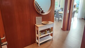 Une armoire en bois, un miroir rond, une petite console blanche ornée d’un vase décoratif et un escalier menant à une salle à manger.