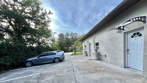 A parking area with a car parked in front of a building with a white door and a window with a decorative grille.