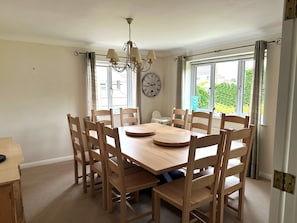 Comedor con mesa y sillas de madera, candelabro, reloj y ventanas grandes con cortinas.