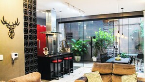 A modern living room with a sofa, a coffee table, and a dining area with a table and chairs. There is a kitchen area with a refrigerator and a sink. The room has large windows and a view of the outside. - Morumbi Triplex House with Jacuzzi (São Paulo)