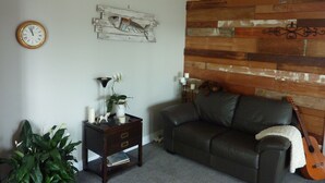 A living room with a wooden wall panel, a clock, a small table with a plant, a couch, and a guitar. - Million Dollar View! (Auckland)