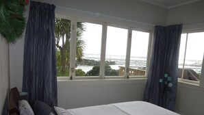 A bedroom with a large window, a bed with pillows, a wooden headboard, and a view of a body of water and buildings. - Million Dollar View! (Auckland)