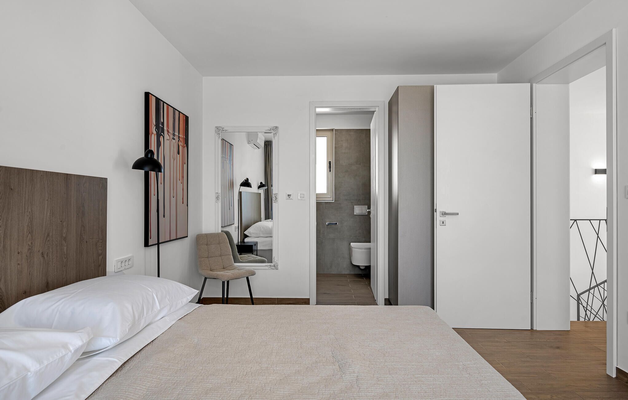 Ein modernes Schlafzimmer mit einem großen Bett, einem Sessel, einem Schreibtisch und einem durchgehenden Türrahmen sichtbaren Badezimmer.
