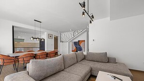 Een moderne woonkamer met een grijze modulaire bank, een eethoek met oranje stoelen en een trap met een geometrisch hekwerk.