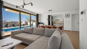 Een moderne woonkamer met een grote grijze modulaire bank, een eethoek met tafel en stoelen, een keuken met moderne apparatuur, en uitzicht op een zwembad en gebouwen.
