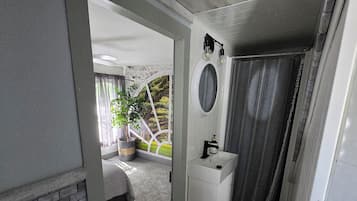 Un baño con un lavamanos blanco, un espejo y una cortina de ducha. Se aprecia una pared de ladrillo y una ventana con vistas a la vegetación.