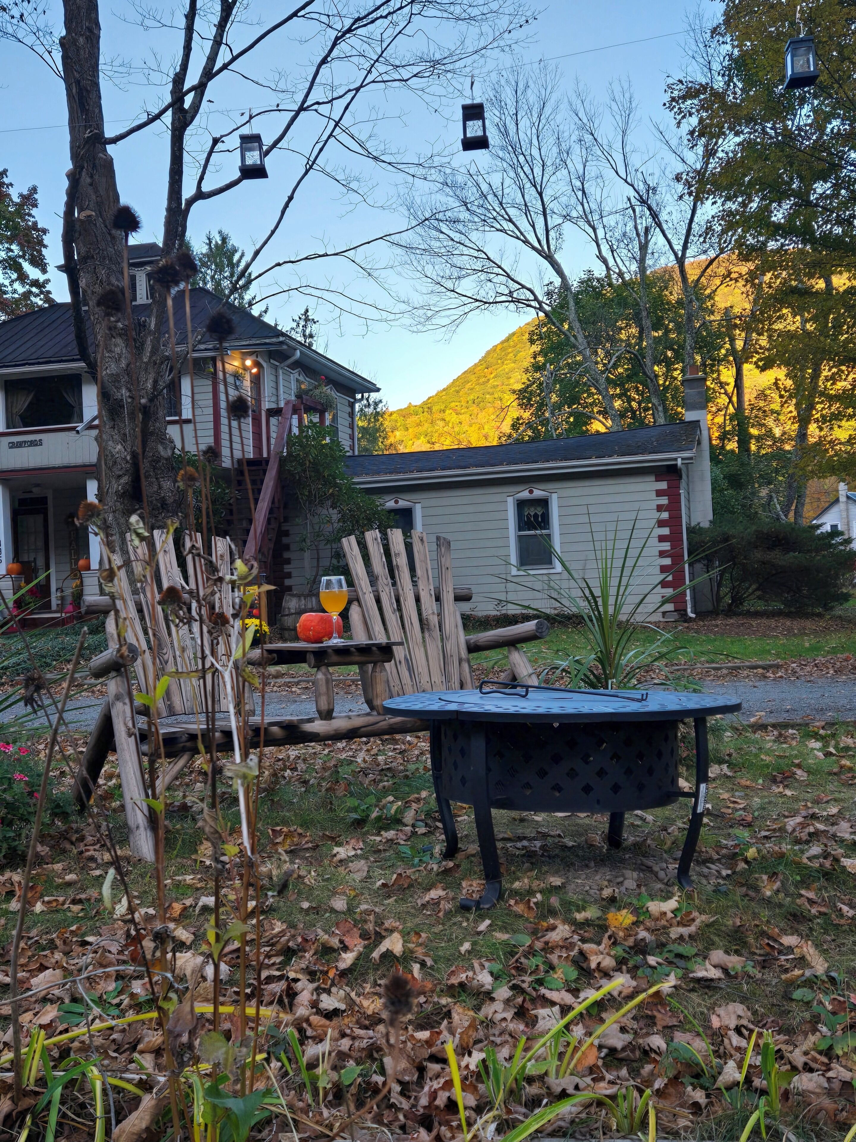 A rustic outdoor seating area with a wooden table and chairs, surrounded by trees and a house with a visible porch.