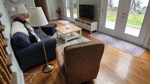 A spacious living room with a large window, a television, a sofa, a coffee table, and a chair. - Peaceful Green Lake Get-A-Way (Ellsworth)