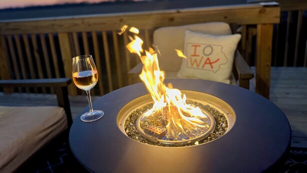 Una chimenea exterior redonda con una copa de vino cerca, sobre una terraza de madera con vista al agua y un cojín con la palabra "IOWA".