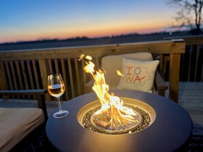 Un foyer extérieur rond, avec un verre de vin à côté, installé sur une terrasse en bois offrant une vue sur l’eau, et un coussin orné du mot « IOWA ».