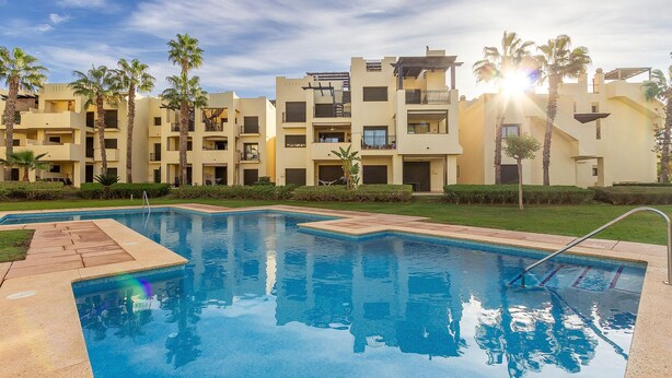 A swimming pool with a clear view of a multi-story building with balconies and palm trees in the background.