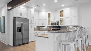 Una cocina moderna con armarios blancos, electrodomésticos de acero inoxidable y una barra con sillas blancas.