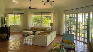 A spacious living room with a large sofa, a coffee table, and a chair. There are large windows with curtains, a ceiling fan, and a decorative light fixture.