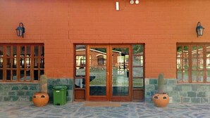 Un bâtiment à la façade en briques, doté de grandes portes vitrées et de deux fenêtres avec des barreaux en bois. Deux plantes en pot sont posées sur le sol.