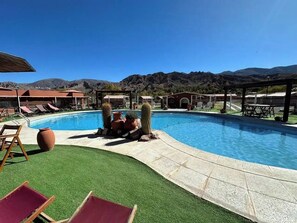 Una piscina con césped verde, sillas de descanso y montañas al fondo.