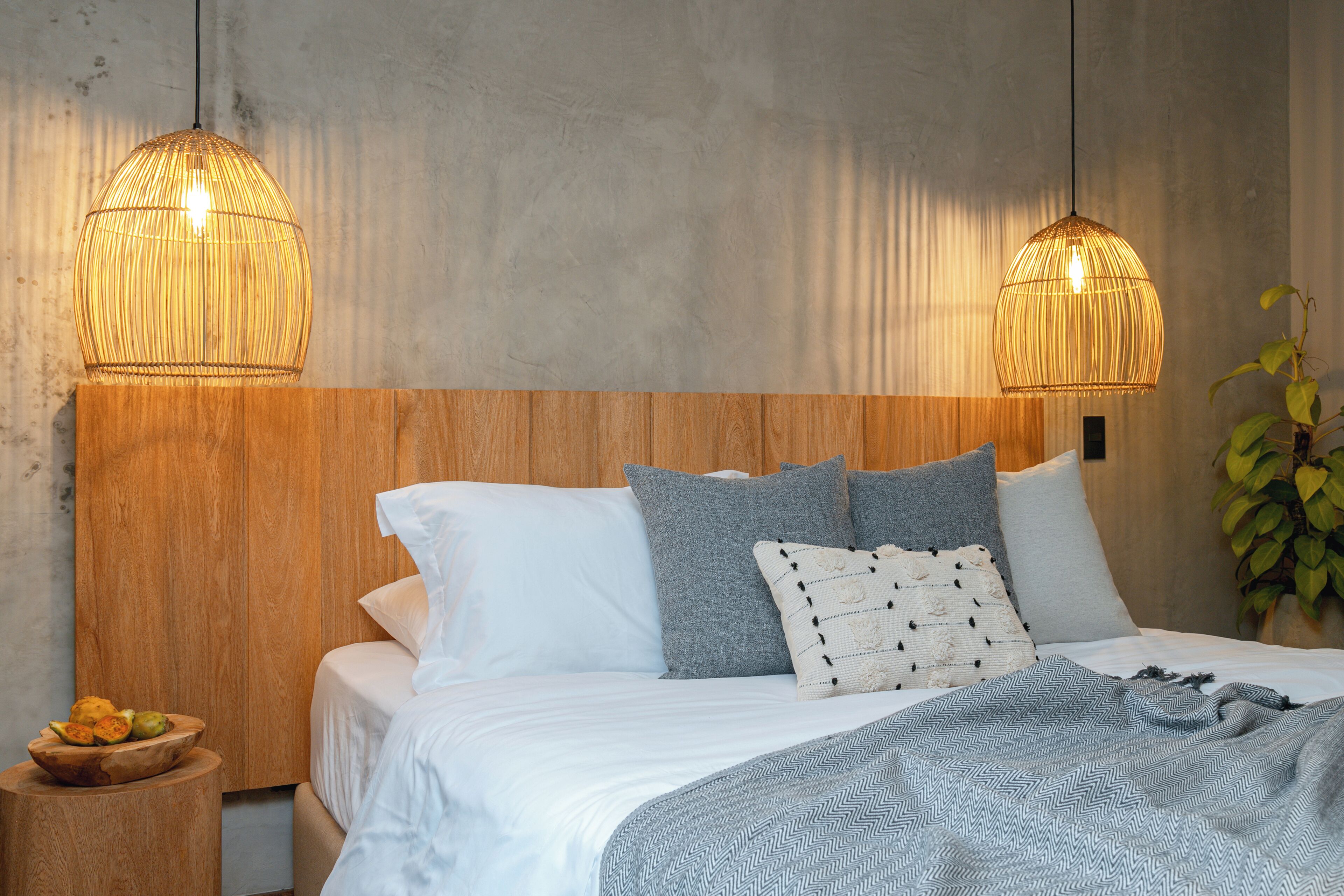 A bedroom with a wooden headboard, a bed with white and grey bedding, two hanging lamps, and a small wooden side table with fruit.