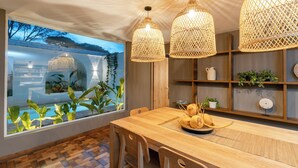 A dining area with a wooden table, wicker pendant lights, and a view of a swimming pool through a large window.