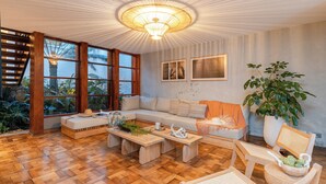 A spacious living room with a large window, a wooden floor, a sofa set, a coffee table, and a potted plant.