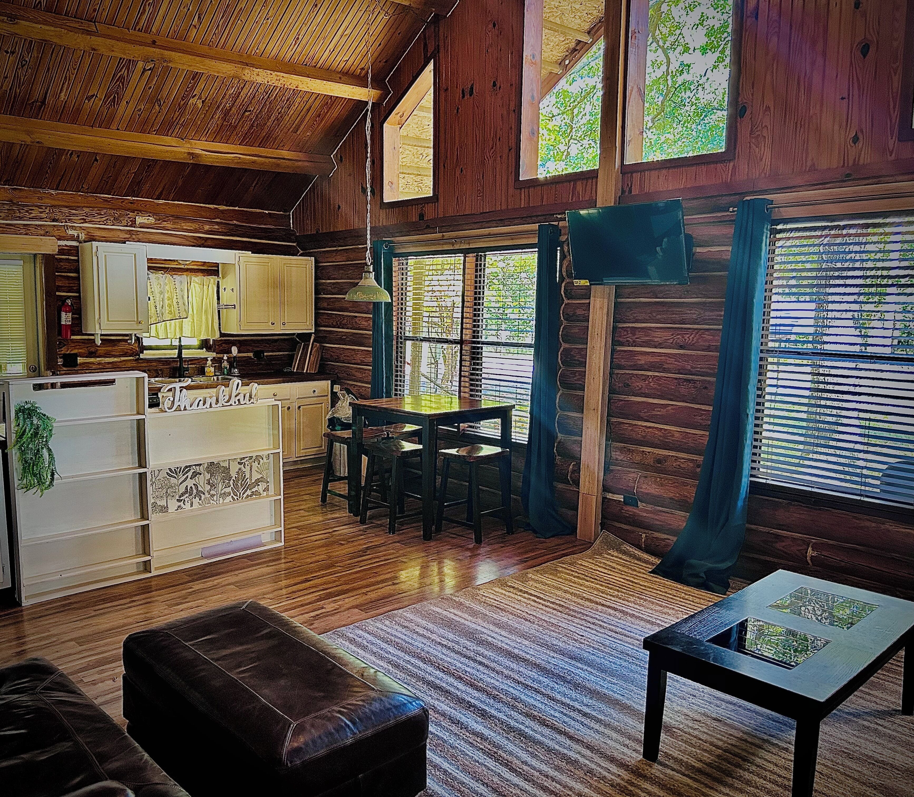 A rustic cabin interior with a wooden floor, a dining table with chairs, a sofa, a small table with a decorative item, and a kitchen area with cabinets and a refrigerator.