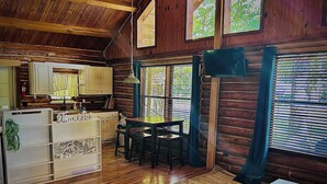 A rustic cabin interior with a wooden floor, a dining table with chairs, a sofa, a small table with a decorative item, and a kitchen area with cabinets and a refrigerator. - Charming 3-bedroom cabin in Eucha with WiFi and FirePit (Eucha)