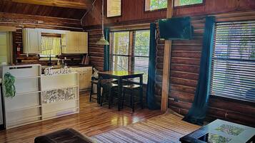 A rustic cabin interior with a wooden floor, a dining table with chairs, a sofa, a small table with a decorative item, and a kitchen area with cabinets and a refrigerator.