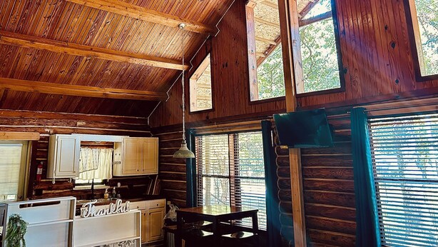 A rustic cabin interior with a wooden ceiling, large windows, a kitchen area, and a dining table. - Charming 3-bedroom cabin in Eucha with WiFi and FirePit (Eucha)