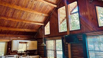 A rustic cabin interior with a wooden ceiling, large windows, a kitchen area, and a dining table.